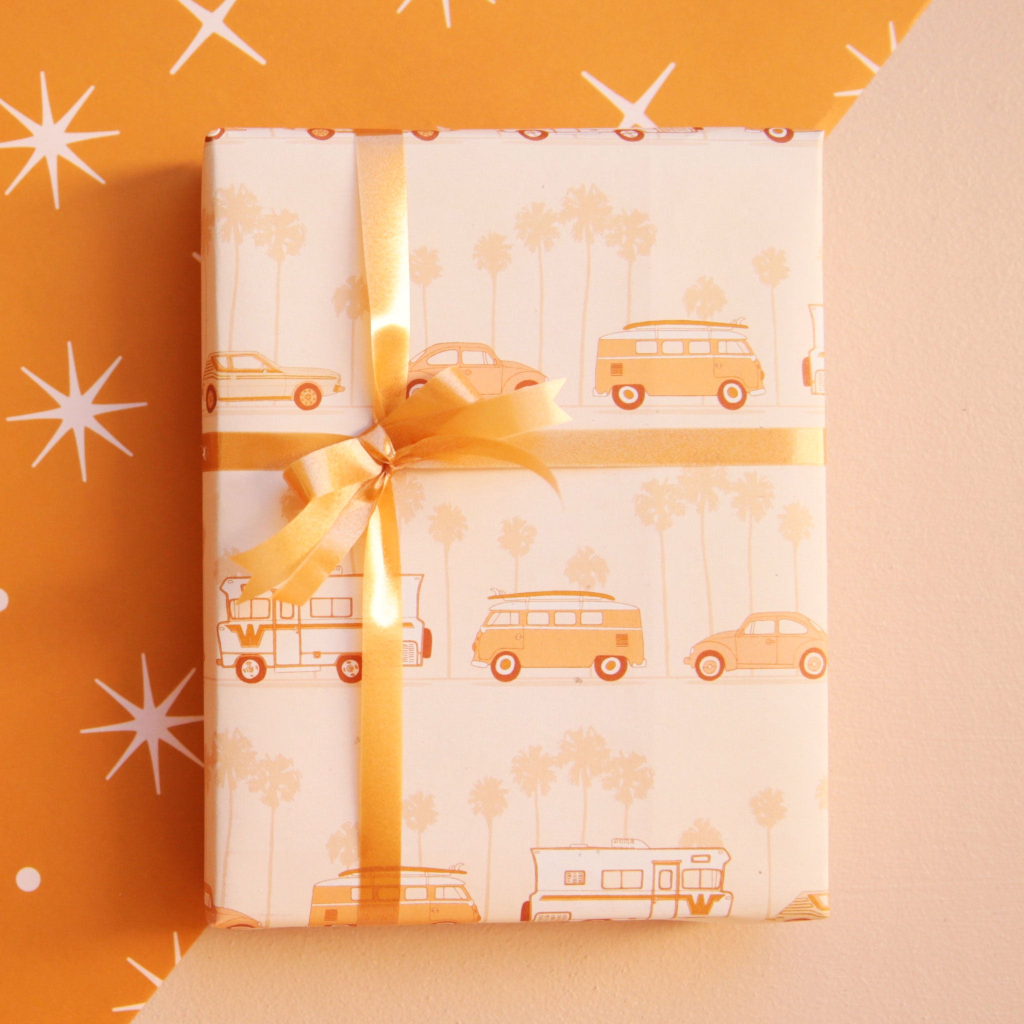 A light orange wrapping paper with a repeating pattern of darker shades of orange vintage VW vehicles including the VW bus, bug and a camper van that are in front of faed palm trees in the background. This photo shows the wrapping paper in use on a gift with an orange ribbon bow.