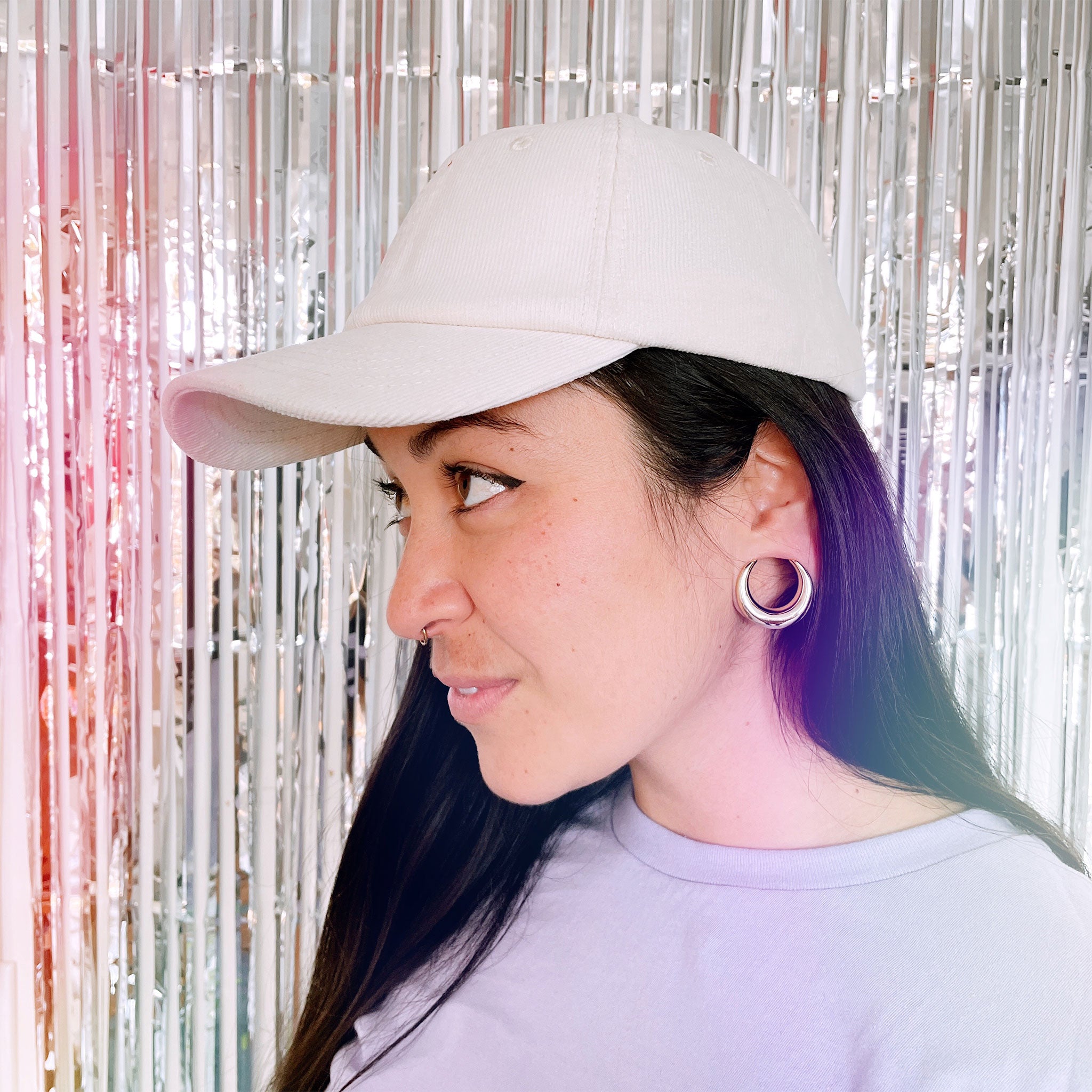 An ivory corduroy baseball hat with a curved bill photographed being worn on a model in front of a silver tinsel background.