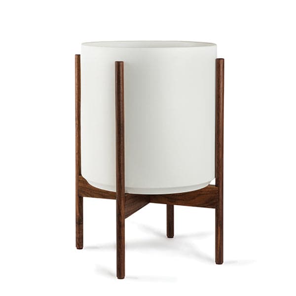 This 5 gallon, cylinder pot is solid white and sits within four spokes of a dark walnut wood plant stand, standing about 7 inches from the ground.