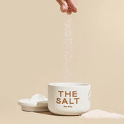 A white ceramic jar with a hand dropping medium grind salt into the jar along with a half moon lid off to the side.