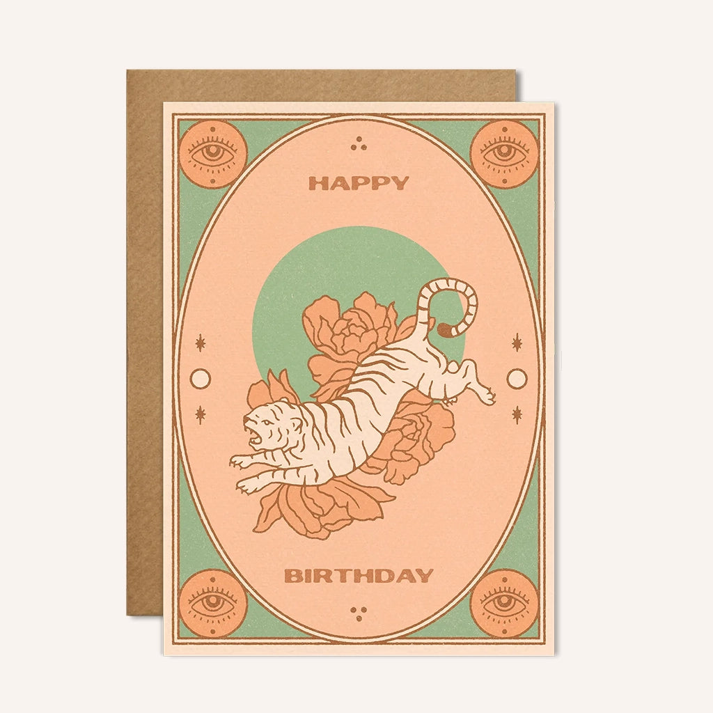 A teal and salmon colored greeting card with a graphic of a cream tiger as well as flowers behind it and seeing eyes in all four corners of the card. The text on the card reads, "Happy Birthday".
