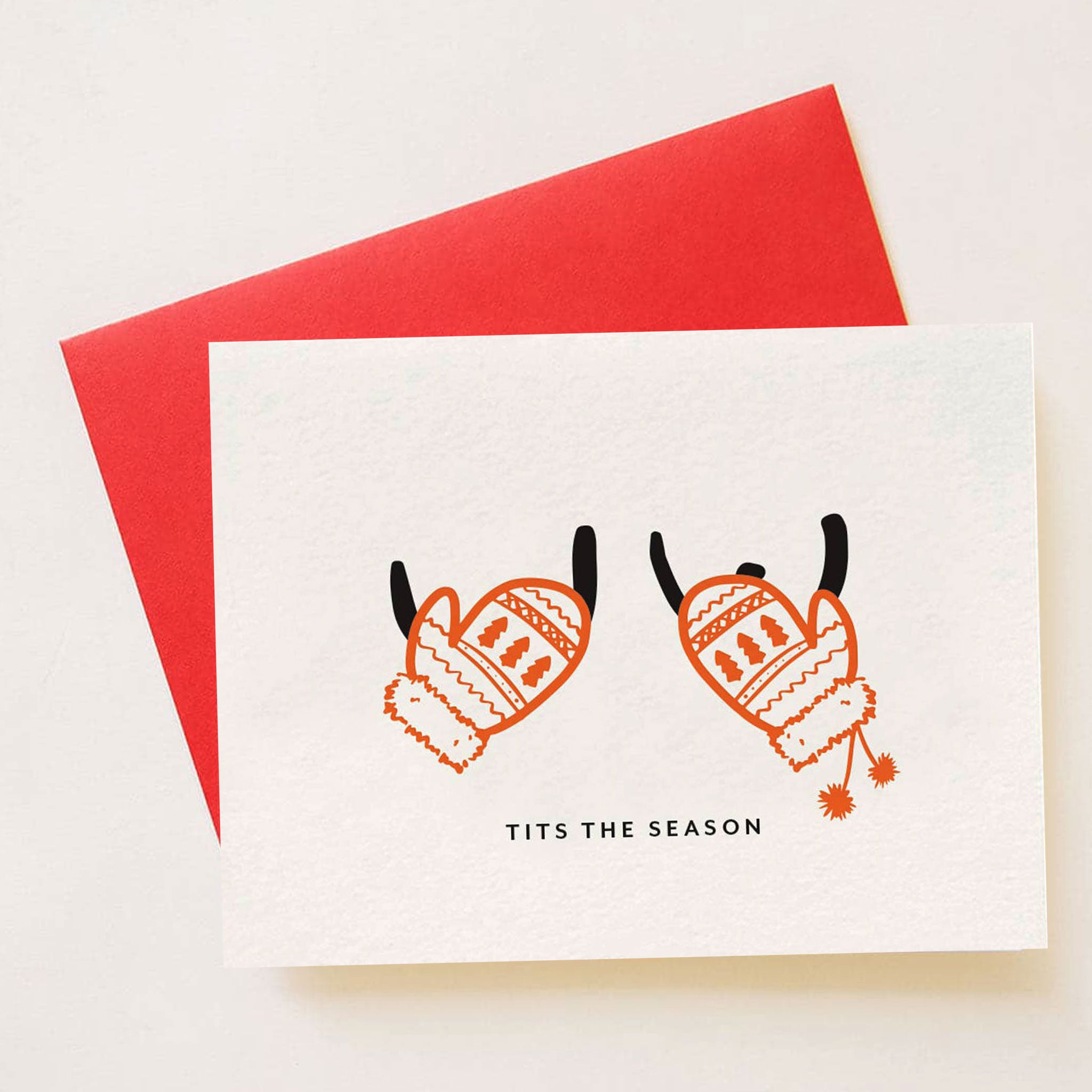 A cream colored card with two outfights of boobs along with red holiday mittens covering them along with text that reads, "Tits The Season". The card is accompanied by a red envelope that is included with purchase. 