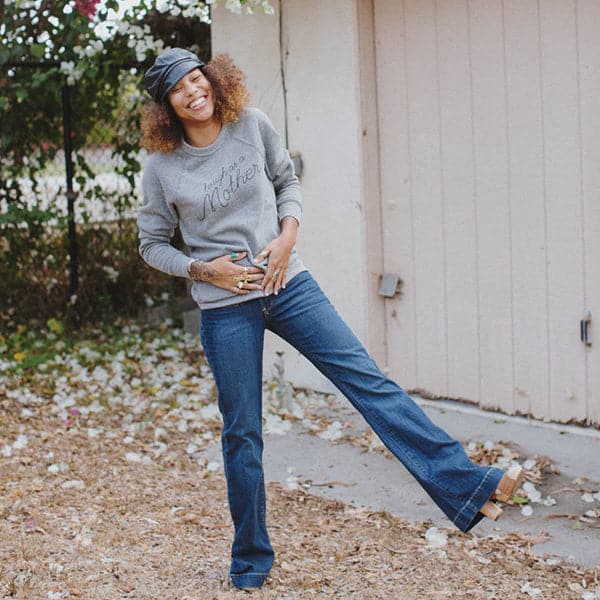 Soft grey screen-printed crewneck reading tough as a mother in black cursive. The crewneck is modeled by a woman with curly hair, a black leather conductor hat and blue jeans.