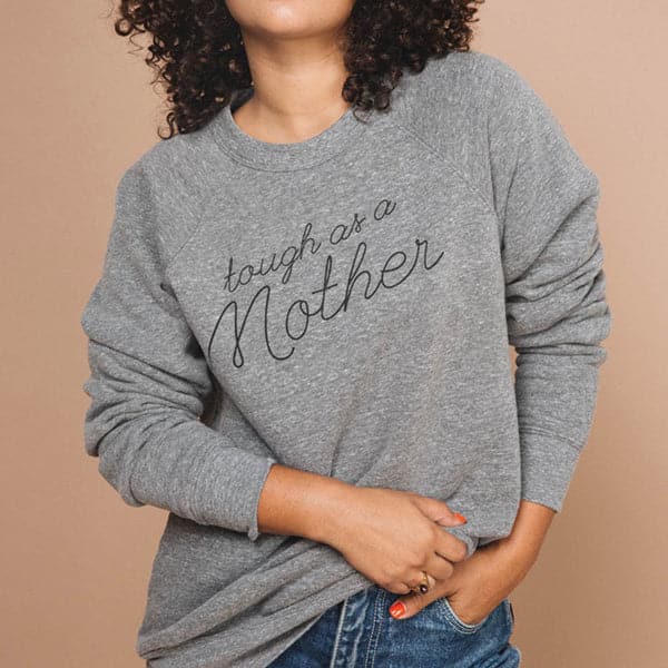 Soft grey screen-printed crewneck reading tough as a mother in black cursive. The crewneck is modeled by a woman with curly dark hair and hand in her pocket.