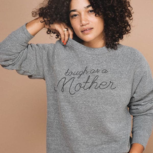 Soft grey screen-printed crewneck reading tough as a mother in black cursive. The crewneck is modeled by a woman with dark curly hair. 
