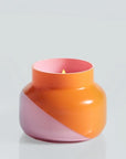 A squatty, round glass jar sits in front of a white background. The jar is split diagonally with the top half being a bright orange color and the bottom half being a soft pink color. The top of the jar is a lot narrower than the rest of the jar. The inside of the jar is the same shade of pink as the outside of the jar. There is a small flame inside the candle. 