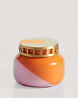 A squatty, round glass jar sits in front of a white background. The jar is split diagonally with the top half being a bright orange color and the bottom half being a soft pink color. The top of the jar is a lot narrower than the rest of the jar. The lid is a light gold color with one row of square cut outs around the side of the lid. There is an orange, rectangular shaped sticker on the left side of the lid. 