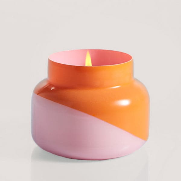 A squatty, round glass jar sits in front of a white background. The jar is split diagonally with the top half being a bright orange color and the bottom half being a soft pink color. The top of the jar is a lot narrower than the rest of the jar. The inside of the jar is the same shade of pink as the outside of the jar. There is a small flame inside the candle. 