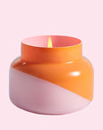 A squatty, round glass jar sits in front of a white background. The jar is split diagonally with the top half being a bright orange color and the bottom half being a soft pink color. The top of the jar is a lot narrower than the rest of the jar. The inside of the jar is the same shade of pink as the outside of the jar. There is a small flame inside the candle. 
