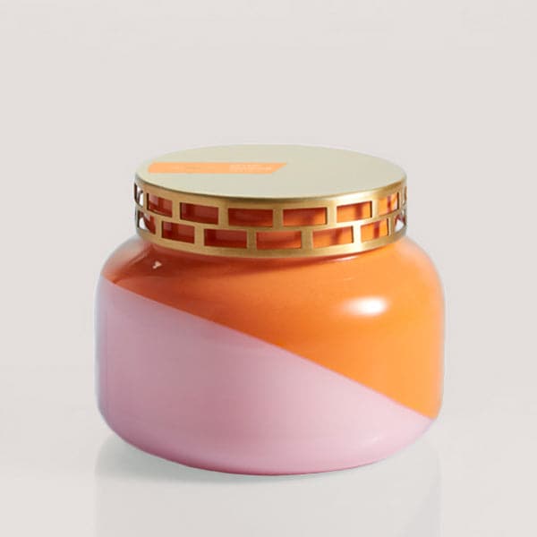 A squatty, round glass jar sits in front of a white background. The jar is split diagonally with the top half being a bright orange color and the bottom half being a soft pink color. The top of the jar is a lot narrower than the rest of the jar. The lid is a light gold color with two rows of rectangle cut outs around the side of the lid. There is an orange, rectangular shaped sticker on the left side of the lid. 