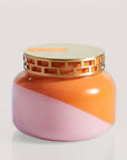 A squatty, round glass jar sits in front of a white background. The jar is split diagonally with the top half being a bright orange color and the bottom half being a soft pink color. The top of the jar is a lot narrower than the rest of the jar. The lid is a light gold color with two rows of rectangle cut outs around the side of the lid. There is an orange, rectangular shaped sticker on the left side of the lid. 