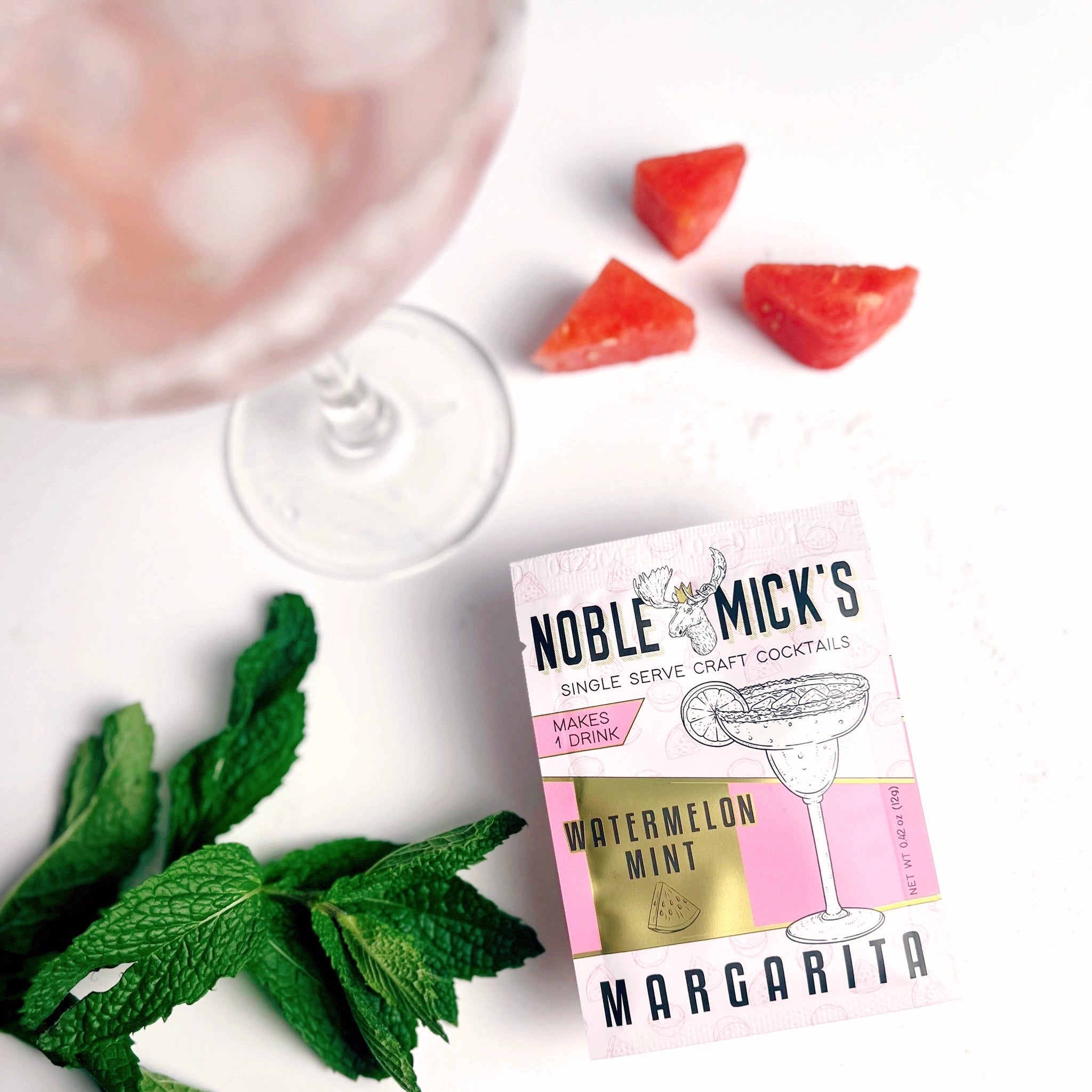 On a white background is a packet of cocktail mix in a white and hot pink packaging that has an illustration of a margarita glass along with black text that reads, "Noble Micks Single Serve Craft Cocktails, Watermelon Mint Margarita" photographed next to a couple slices of watermelon, mint and a cocktail glass with pink liquid inside. 