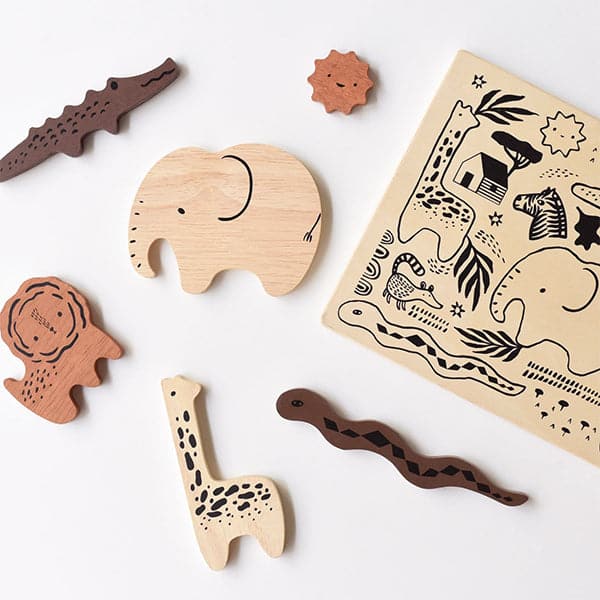 6 wooden puzzle pieces shaped as African animals including a beech wood elephant and giraffe, teak colored lion and chocolate crocodile and snake. The pieces lay besides an ivory block puzzle tray with smooth rounded edges and black detailing throughout. The puzzle lays against a solid white background.