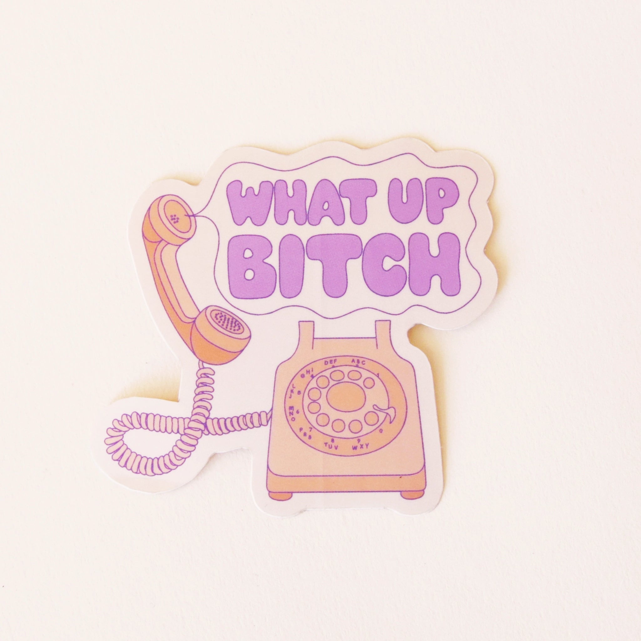 On a cream background is a vinyl sticker of an old school pink phone with a speech bubble coming out of the phone that reads, &quot;What Up Bitch&quot; in purple letters. 