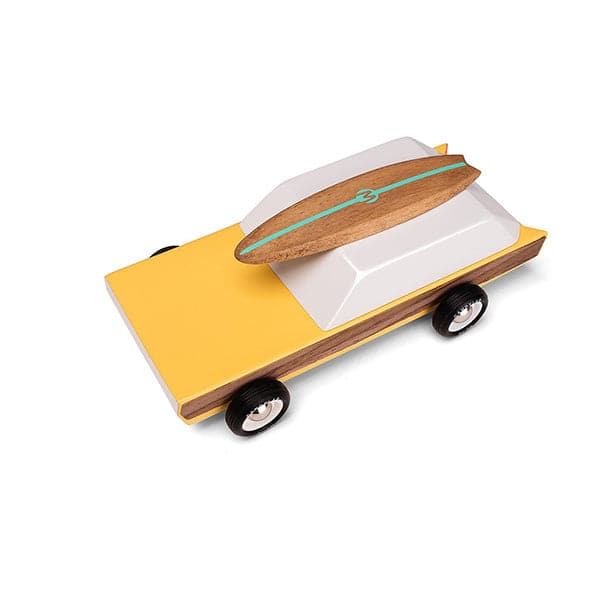 Birds eye view of wooden station wagon model with elongated daffodil yellow body with walnut veneer-paneled sides. Wheels are jet-black with white and silver rims. The windowed top half consists of a single white trapezoid shaped wooden block. A magnetic beech wood surfboard lays directly on top with a sea foam stripe a &#39;M&#39; logo. 