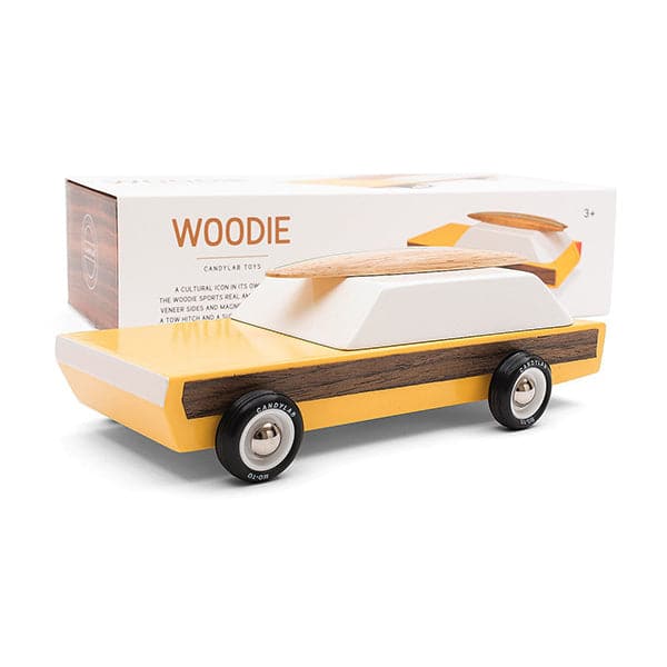This wooden station wagon model has an elongated daffodil yellow body with walnut veneer-paneled sides. Wheels are jet black reading &#39;Candylab Mo-To&#39; around white and silver rims. The windowed top half consists of a single white trapezoid shaped wooden block. A magnetic beech wood surfboard lays directly on top. A white packaging box is propped behind displaying &#39;Woodie&#39; in orange lettering and a picture of the model itself. 