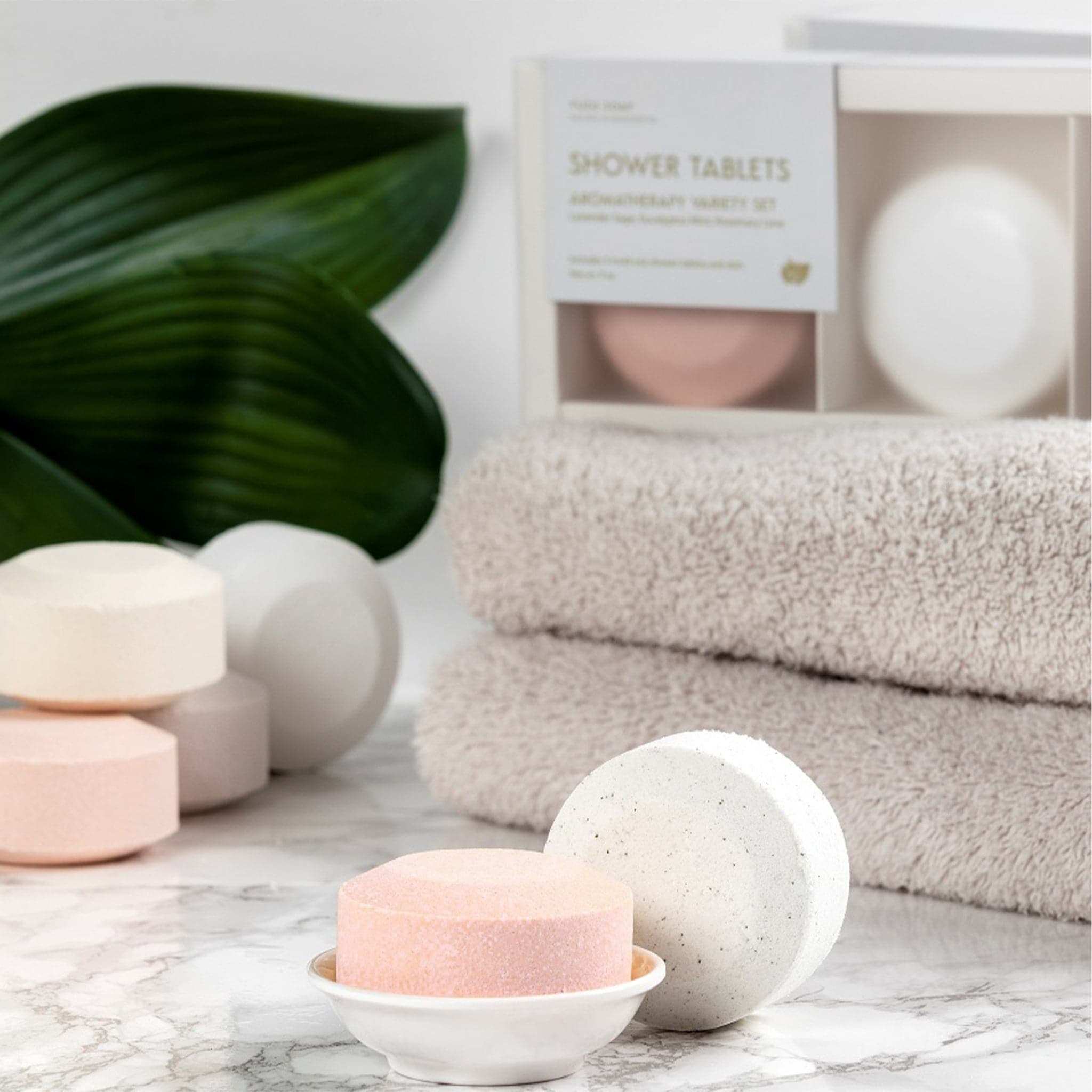 Multiple white and pink shower tablets on a marble bathroom counter with natural colored towels and the dark leaves of a plant. The shower tablets are about palm sized.